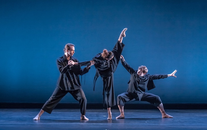 Three dancers in blue baggy costumes arrange themselves near one another. A woman in the center looks under her leg that kicks high into the air. A man crouches underneath her leg while another supports her balance by holding her arm.