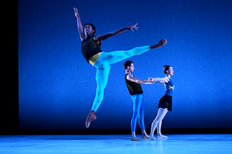 A man jumps in a springy first arabesque while two couples holding hands are seen in the background