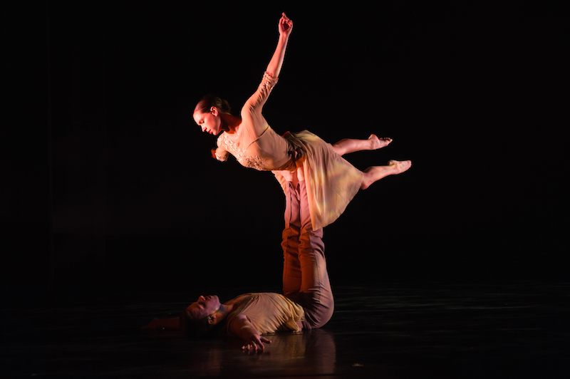 A man lays on his back with his feet extended and supports his partner above him. The female dancer, in a costumed leotard and skirts appears to be floating in air save for the fact her partner is supporting her by his feet.