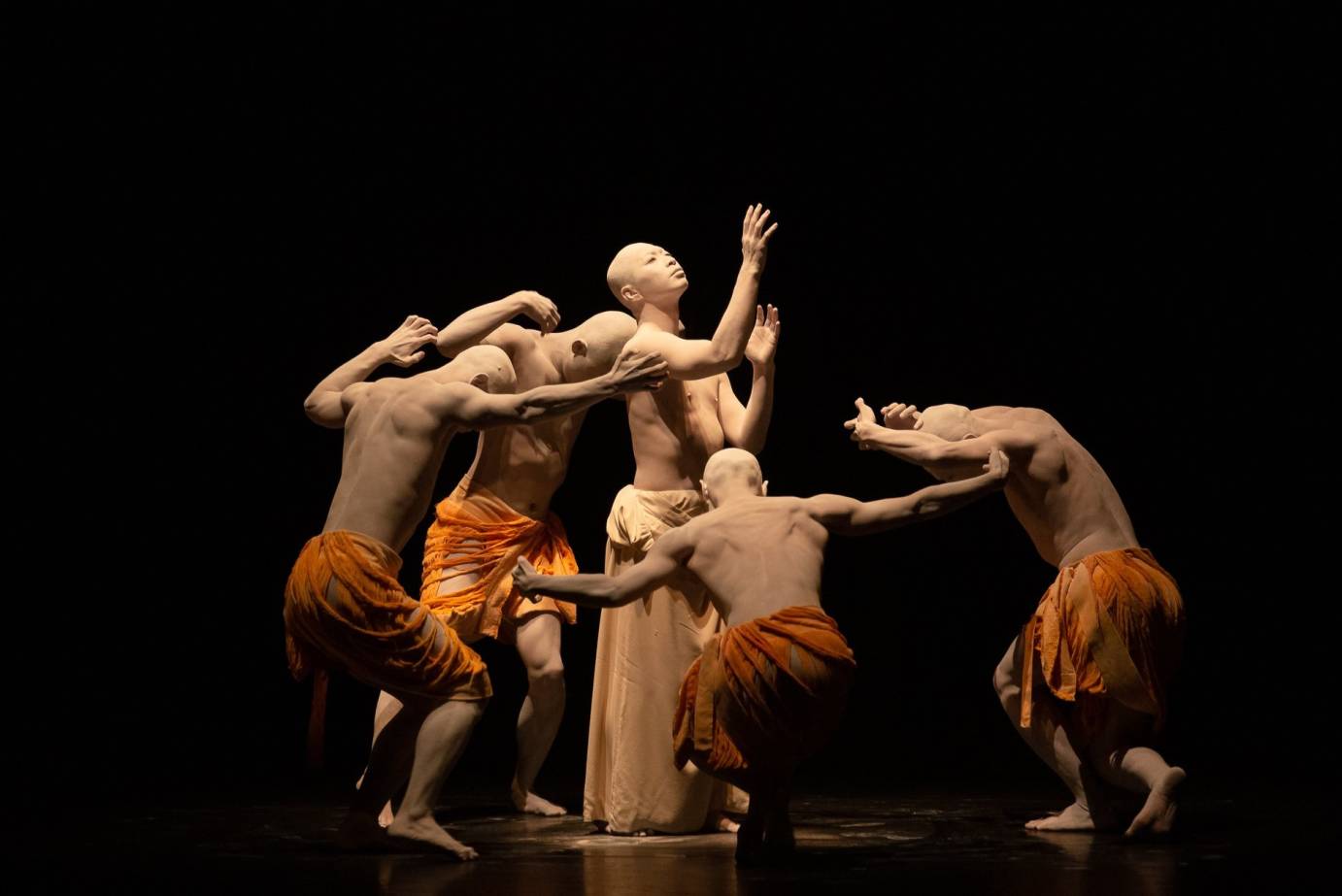 4 dancers clothed in orange cloth around their waste circle around a dancer who is reaching upwards