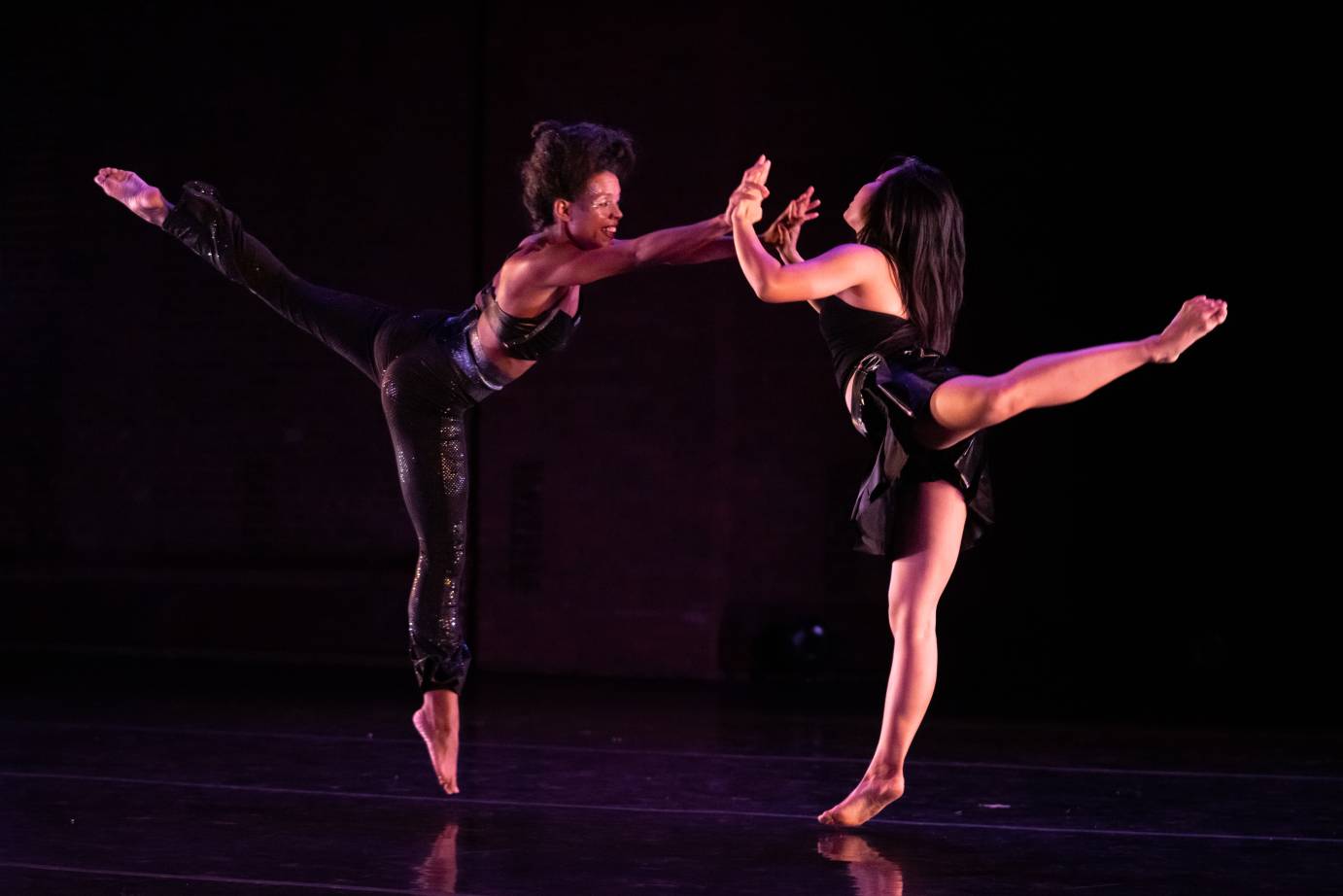 2 female dancers holding hands in a jump with one leg extended behind them