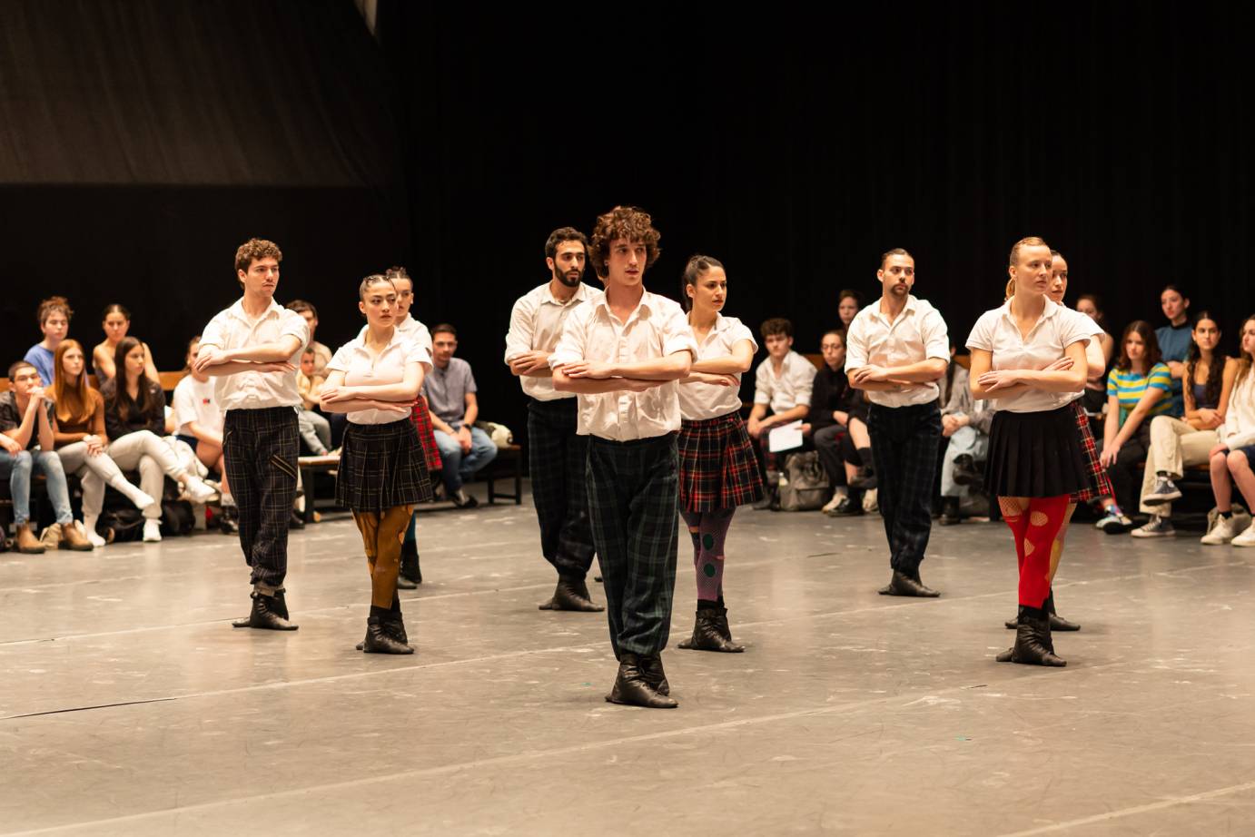 9 dancers standing in formation, in 5th position with their arms crossed at their chests