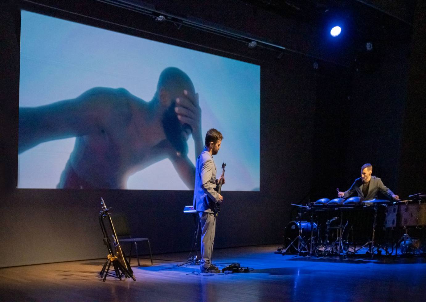 guitarist and drummer onstage in front of projection where dancers stands holding his face in his hand, we can only see his upper torso and head