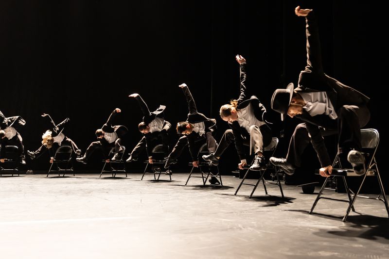 7 dancers pushing themselves up out of a chair, both feet off the floor one hand upraised the other pushing on chair