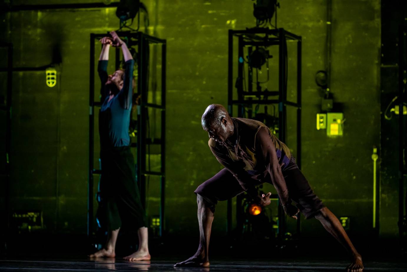 male dance in a low squat reaching to floor. Dancer behind and to the left with arms stretched up