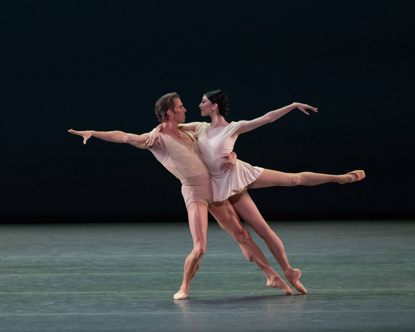Duet. Female and male dancer facing each other, she is leaning into him in an arabesque, his free arm reaching behind him