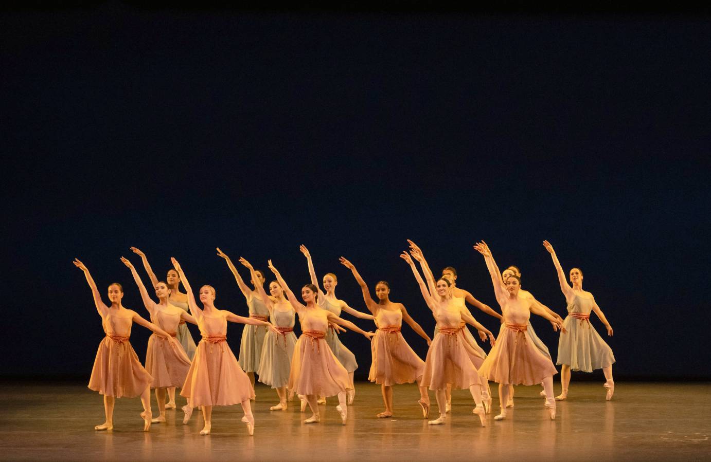14 female dancers in tight formation in tendu with one arm up, one arm to side