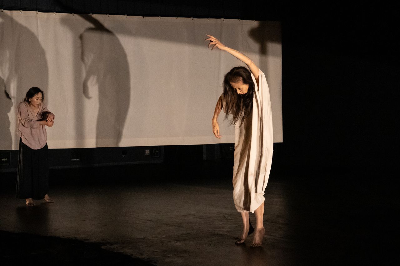 female dancer in white nightgown stands in a contraction with one arm above her head. another female dancer in backgroung