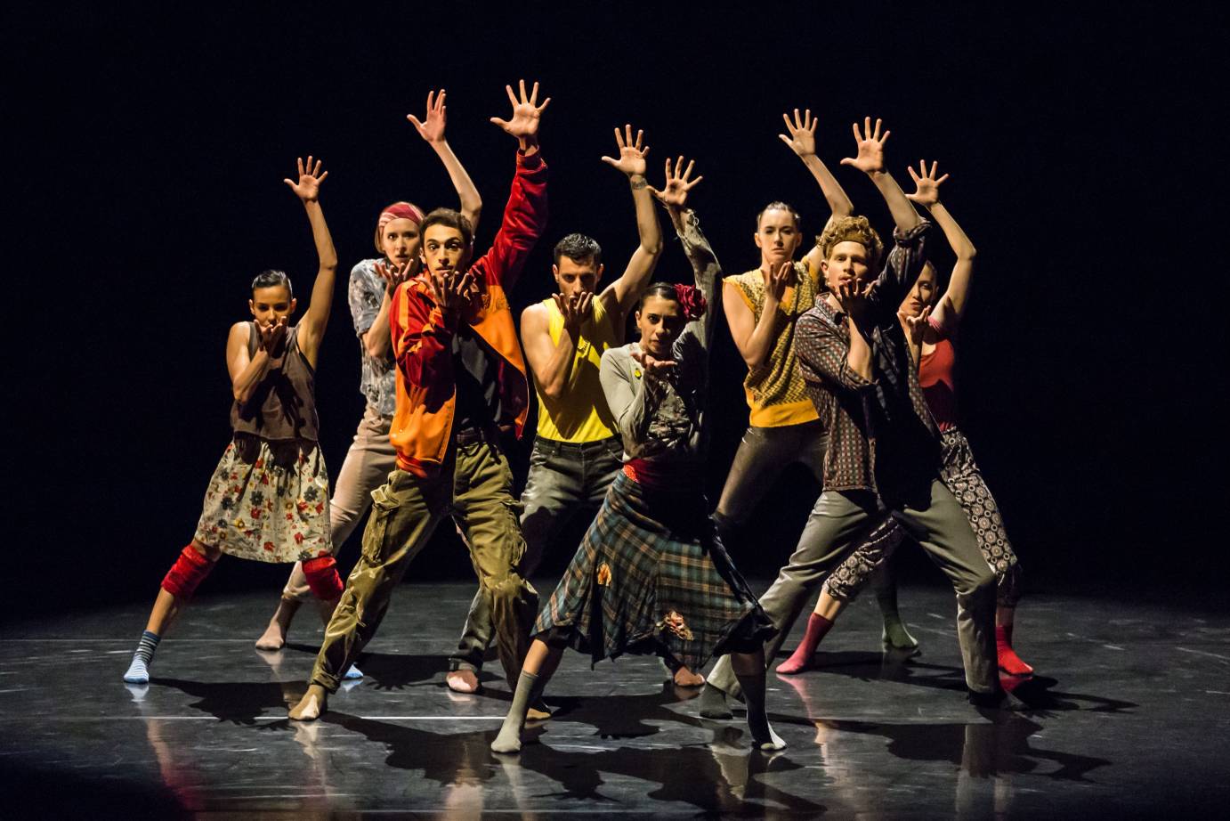 8 dancers in a clump, one arm extended up and fingers splayed