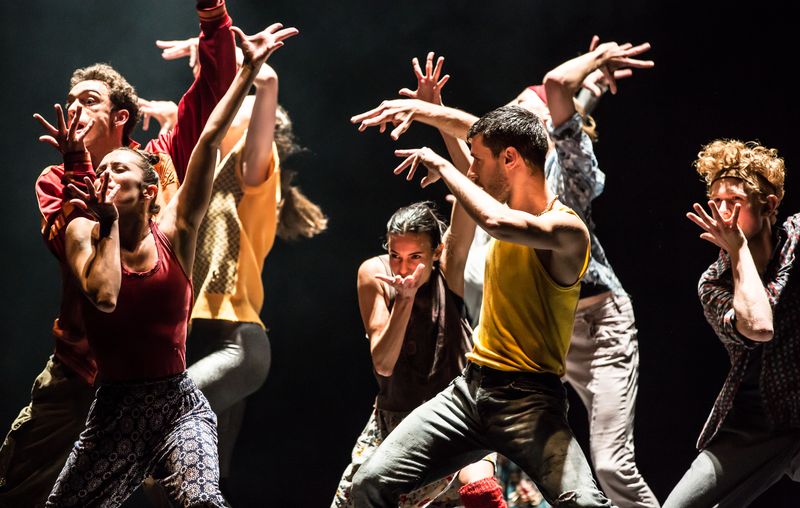 8 dancers in a clump in various shapes, one hand up and one hand in front of mouth, fingers splayed