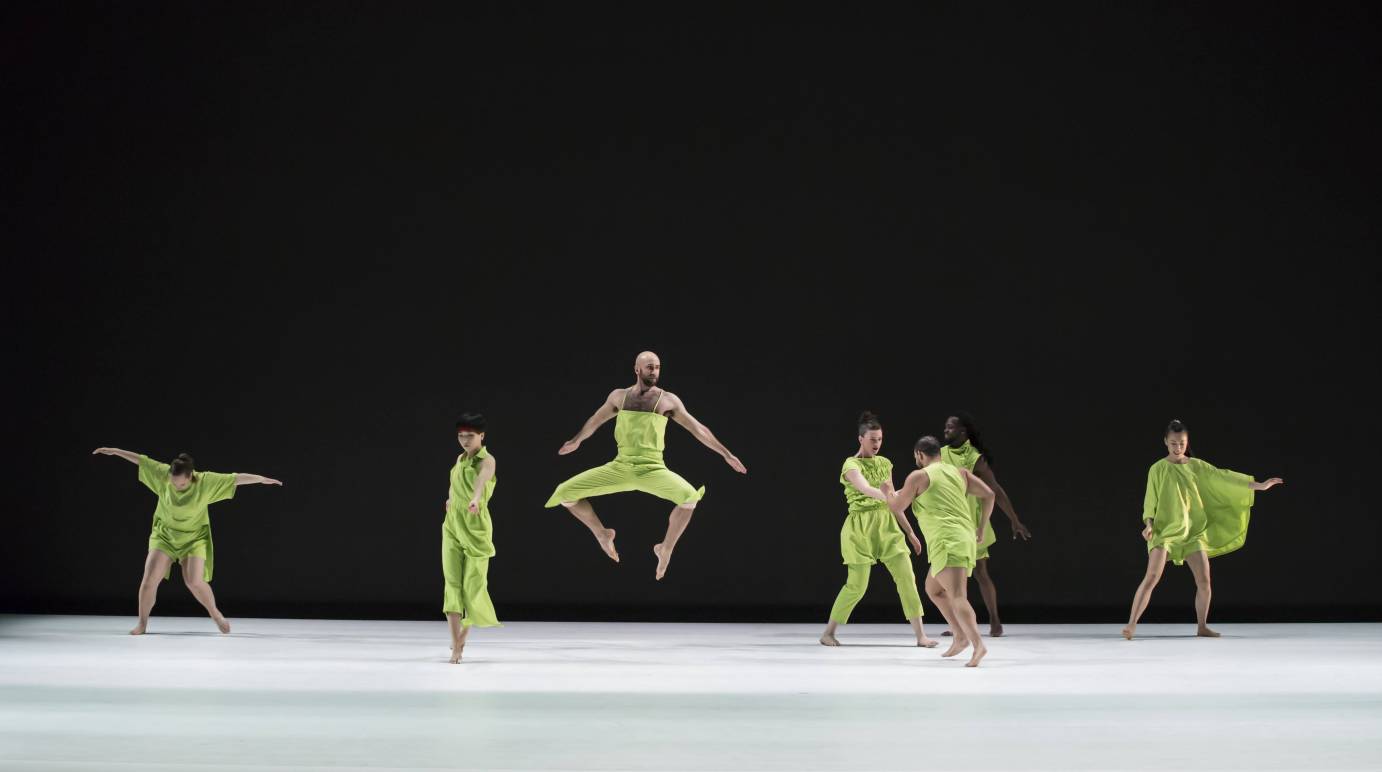 7 dancers on stage. Dancer in center jumping with both legs bent underneath his body