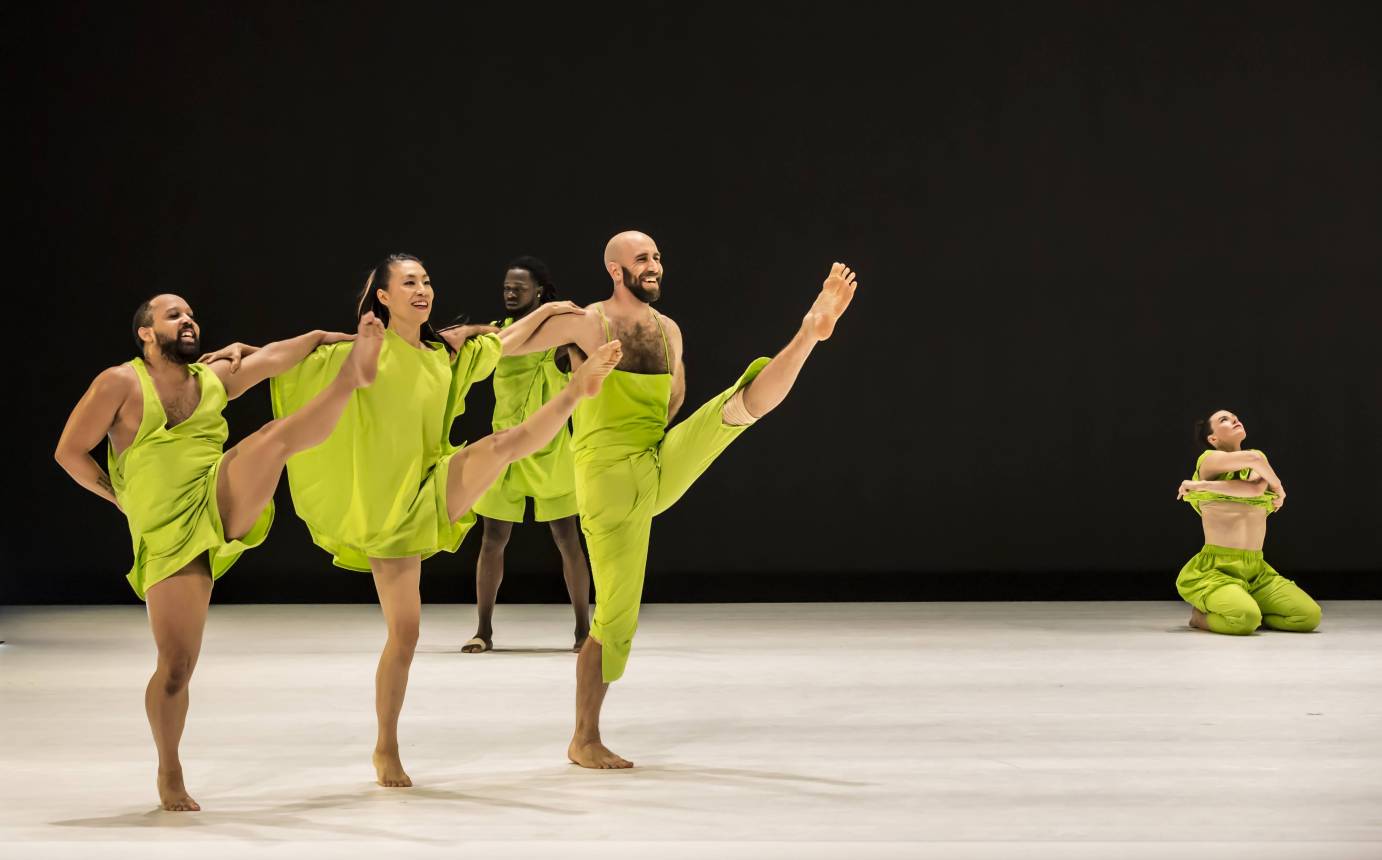 3 dancers holding shoulders and kicking left leg. One dancer kneeling in background