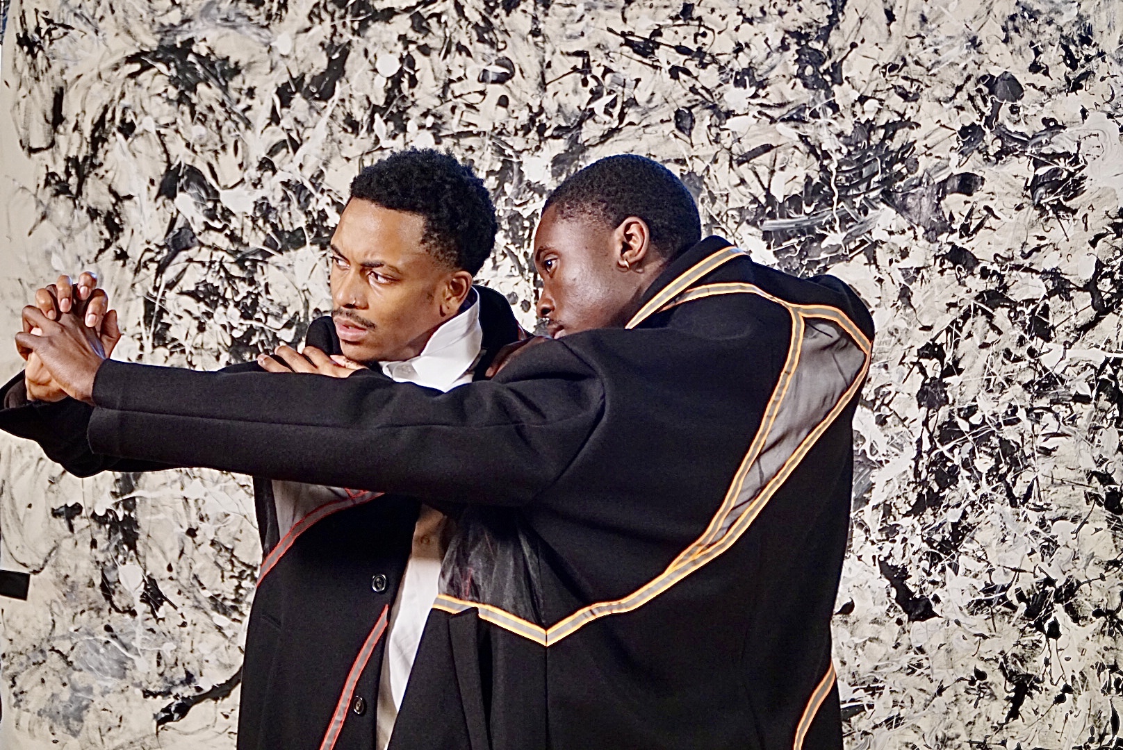 two black men in black ceremonial jackets hold on to one another  against a huge black and white splattered canvasBallet X