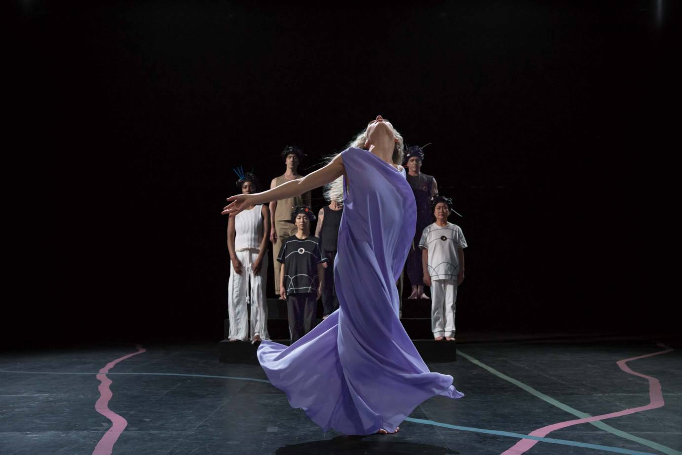 A female dancer clad in a purple dress sashays before a crowd of other dancers who stand watching her.