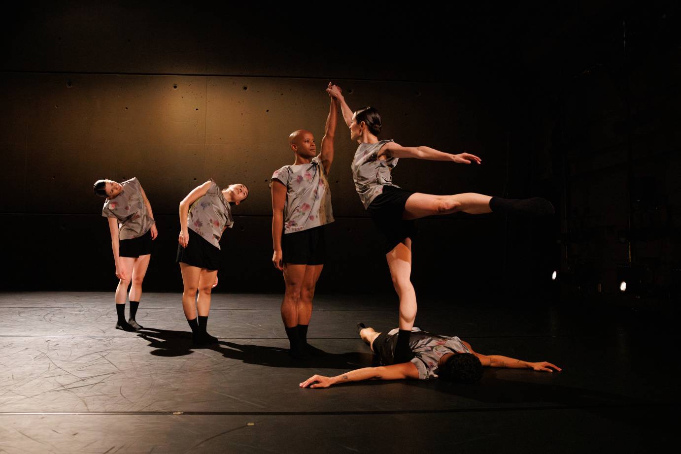 five dancers in black socks, black shirts, and gray tshirts with splotched designs on them create a line formation in which three dancers face one dancer in an arabesque, who happens to be standing on the back of another dancer 