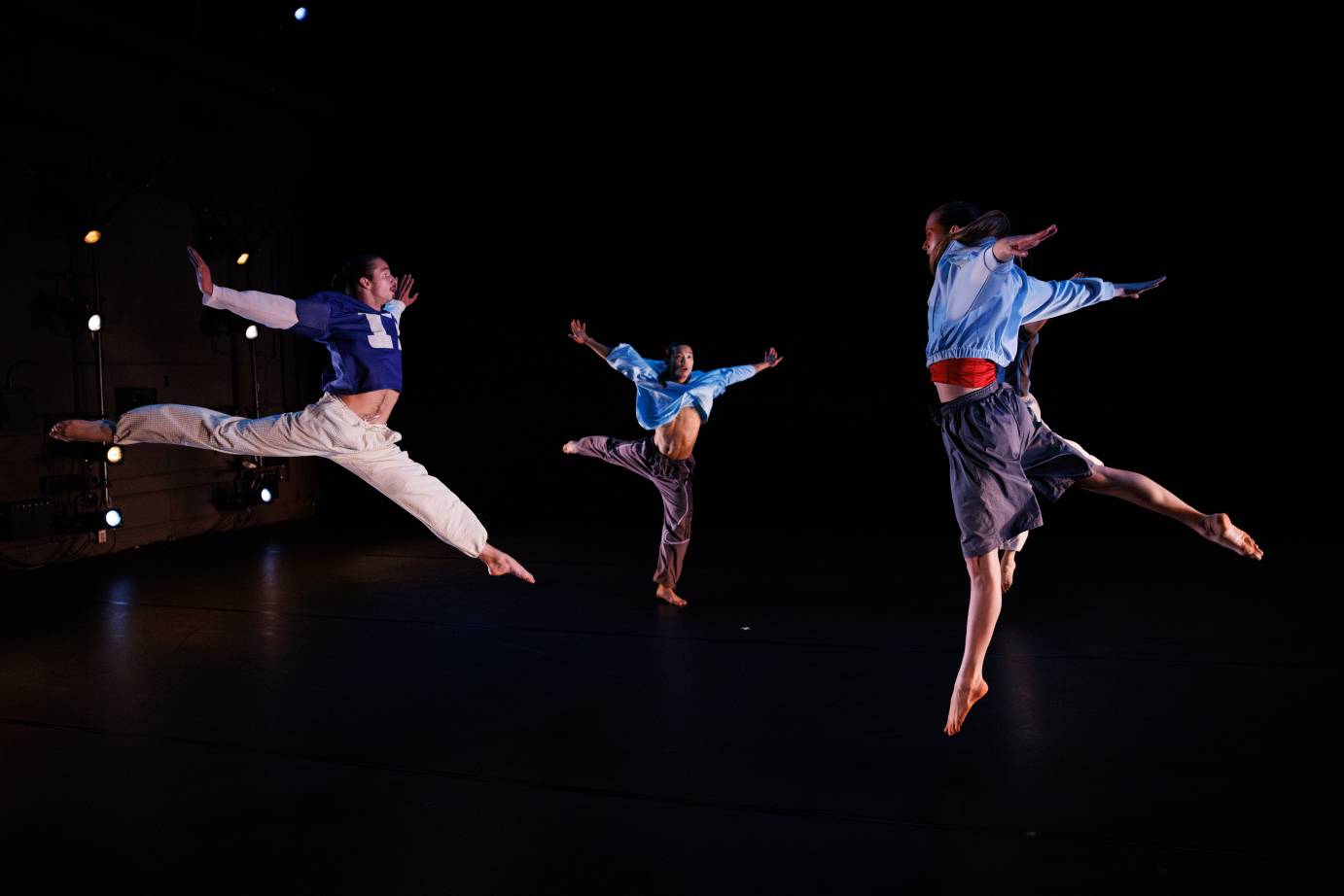 three dancers in colorful blues, whites, greys, and reds, arabesque jump towards eachother on stage