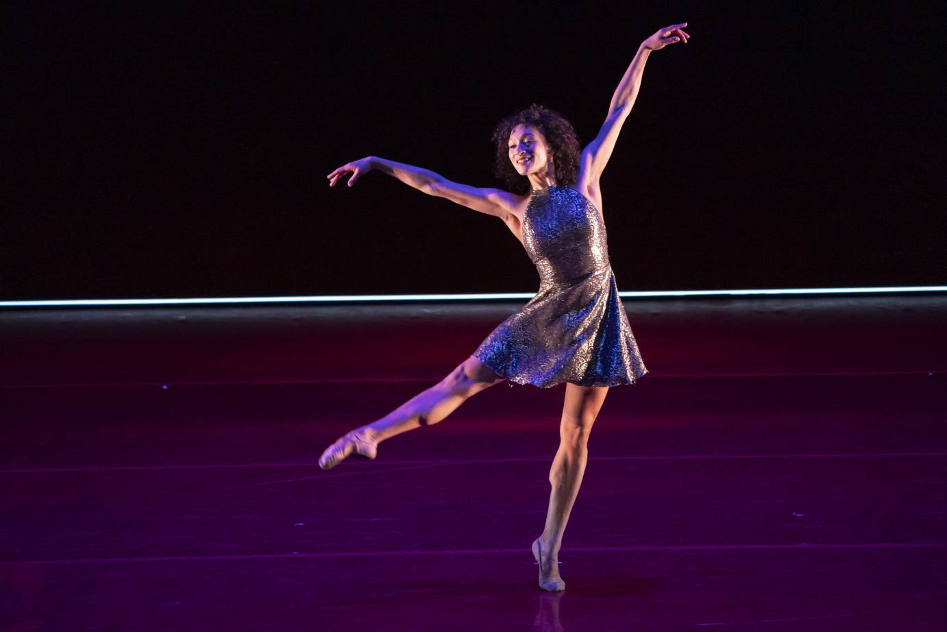A woman in a silver dress stretches upward on tip toe, arms tossed on the diagonal, with a smile on her face.
