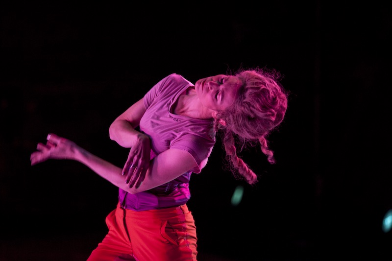 Elizabeth Dement bends her body to the side. Her skin and hair are dyed hot pink. She wears red pants.