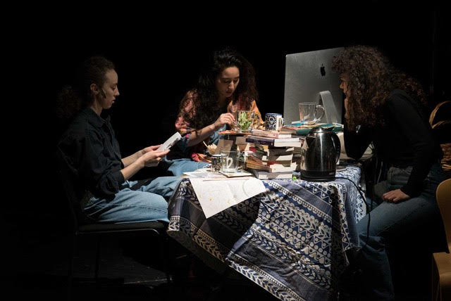 Three artists sit around a table. They are in discussion. Mugs, paper and books litter the table.