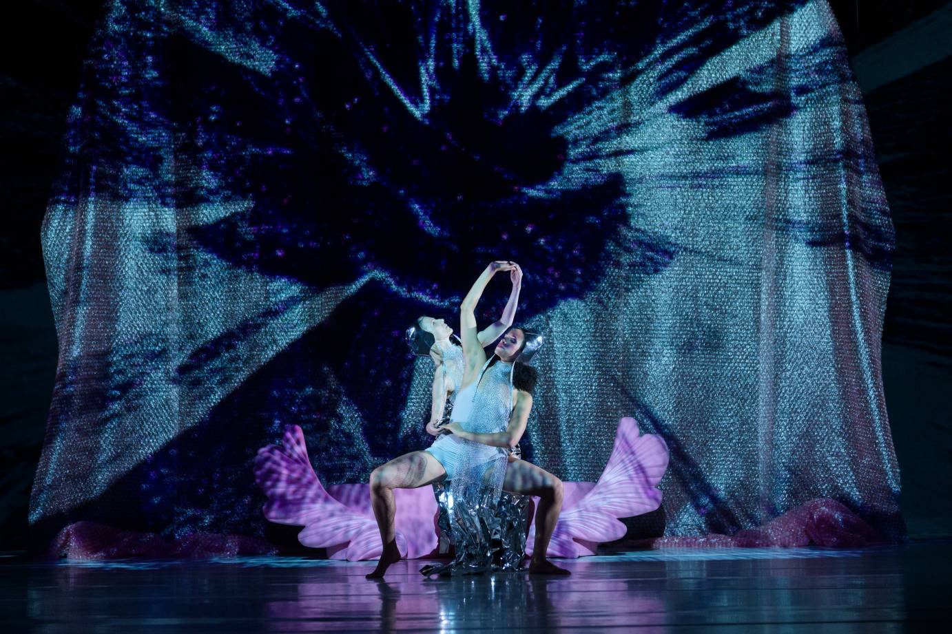 Two women create a garland with their raised arms while in a deep second position plie