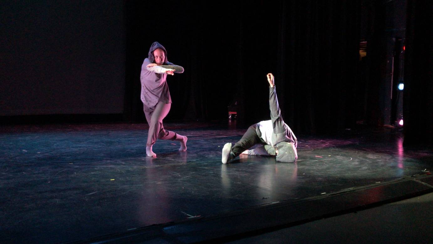 Two dancers in sweatsuits perform on stage. One is in a pose on the floor while the other extend's their arm out towards the audience.