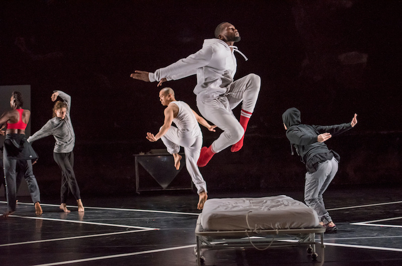 The dancer in light grey leaps by a bed; his red-socked feet tuck underneath him. Other dancers leaping and posing are in the background.