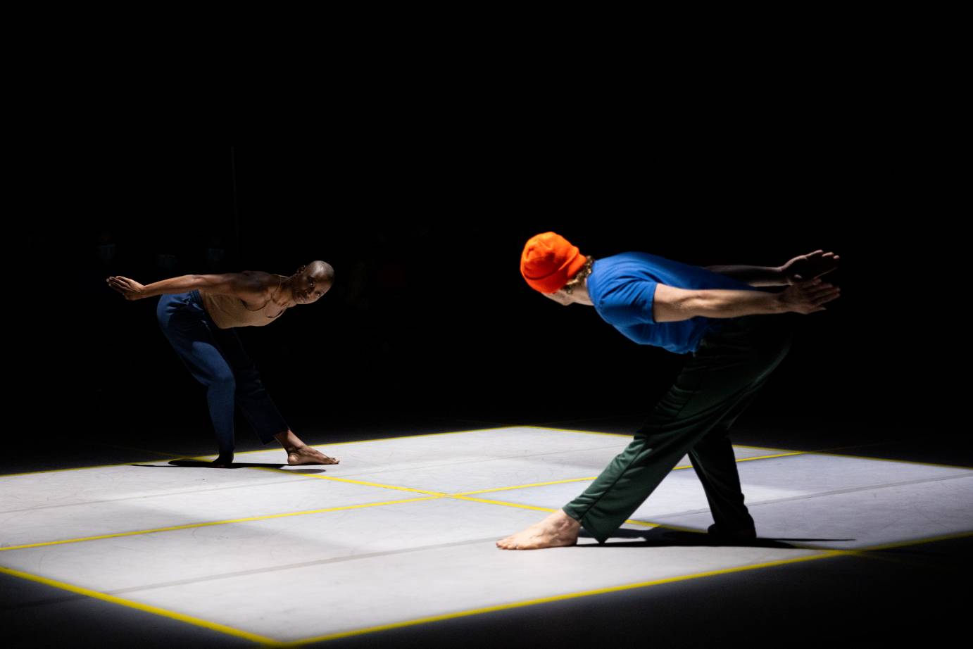 two dancers, a black woman in blue pants and a light brown top and a fair skinned man in a bright blue top, dark pants, and orange cap, face each other mirroring each other. Their torsos are parallel to the floor, the lean towards their right leg as the left leg bends, their arms are extended behind them. 