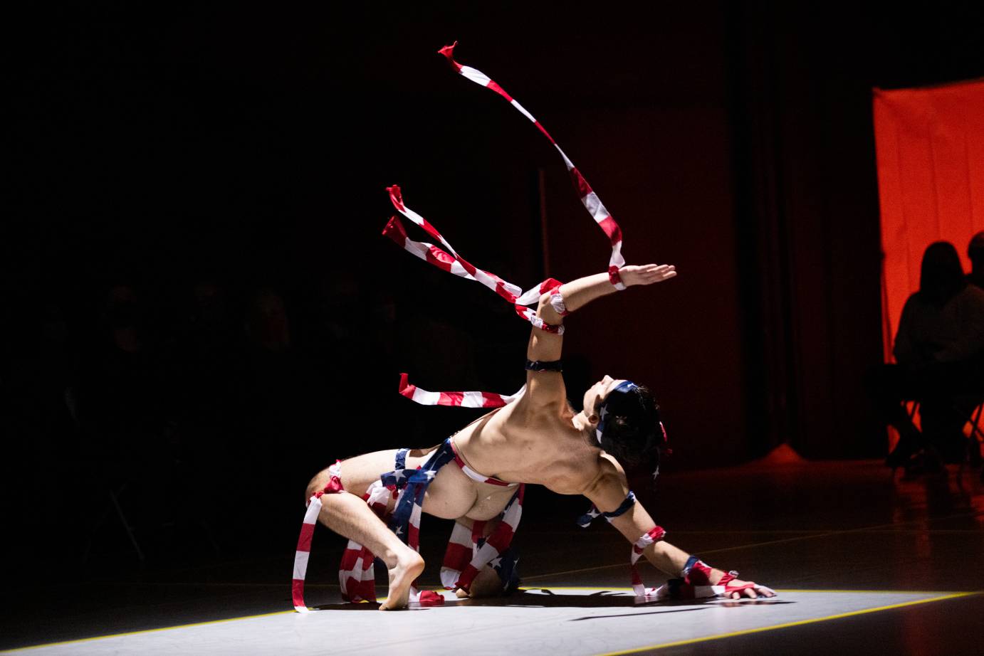 a fair skinned man dressed in scraps of the American Flag and nothing else, falls back ward onto one arm as his other arm reaches upward, rags flying in the air. 