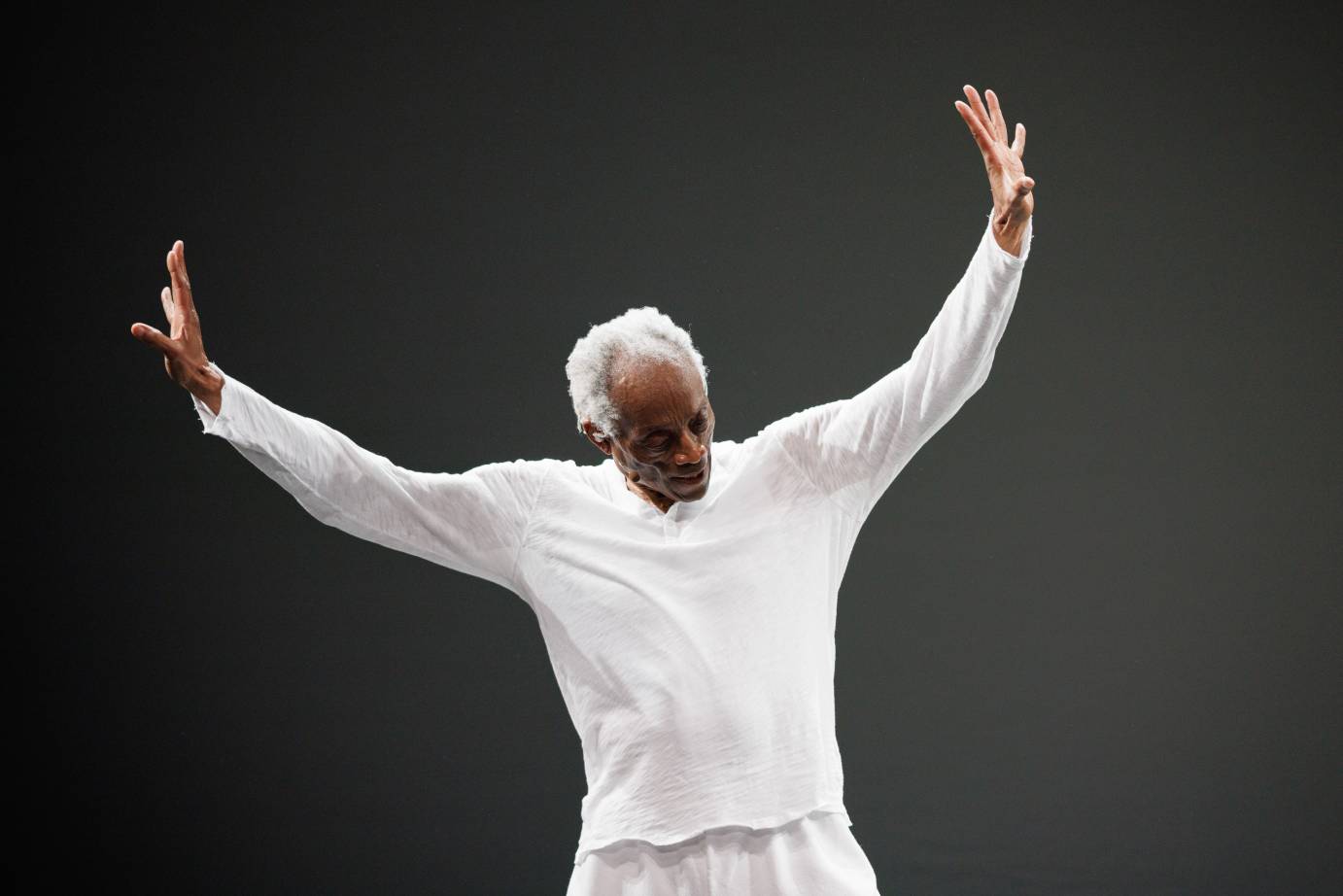 A white-haired, white-costumed man tips head down while both arms are lifted to the diagonal palms out.