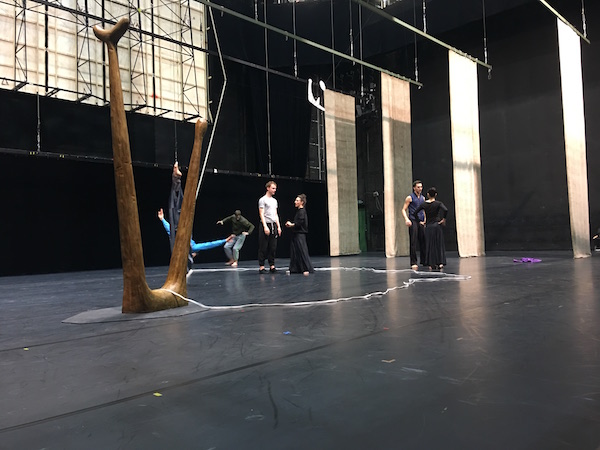 Dancers in rehearsal clothes practice on stage by a long wooden set piece.