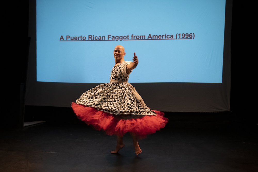 Arthur Aviles of BAAD in the Bronx performs a solo in a checkered dress with a full skirt accented with red flounce at the bottom -- on screen behind him we read 