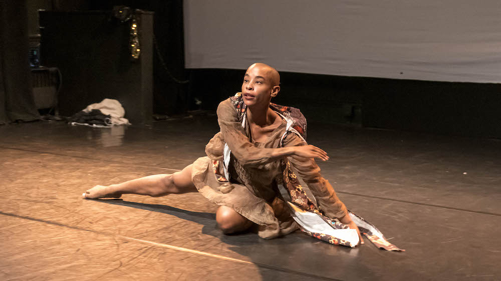 a bald pated, black dancer sits on a bent knee with their other leg extended to the side on the floor, they wear a brown outfit and seem to be looking at the audience as they gesture with a bent arm in front of tehm