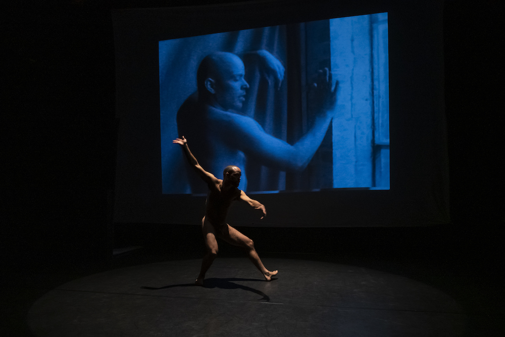 barely lit we appreciate the body of a man in motion behind him projected in bluish light is a film of the original dancer and choreographer Arthur Aviles