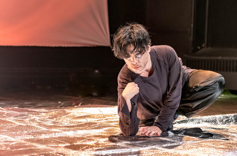 an Asian man with mussed black hair and white markings under his eyes leans on his elbow on a blanket of silver..
