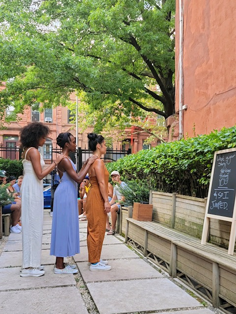 Three women of color, artists of the Blue Morph Collective, wearing pantsuits in white, periwinkle, and orange-brown stand in a line outside of The Garden of Hope in BedStuy, Brooklyn. They seem to be meditating, while staring at the green leaves in front of them, while onlookers observe.