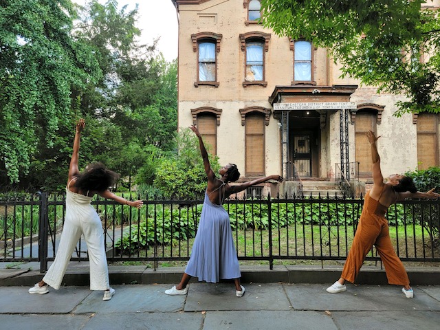 Three women of color in pantsuits of white, periwinkle, and orange brown with white sneakers, stand with their backs facing us arms outstretched while in front of the gates of the Order of Tents brownstone in Bed Stuy, Brooklyn