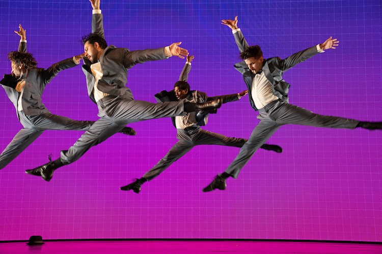 against a pink purple background for men wearing grey suits, white shirts, and no ties, leap into the air, their legs making a wide v shape. the last man in the group dangles a cigarette from his mouth.