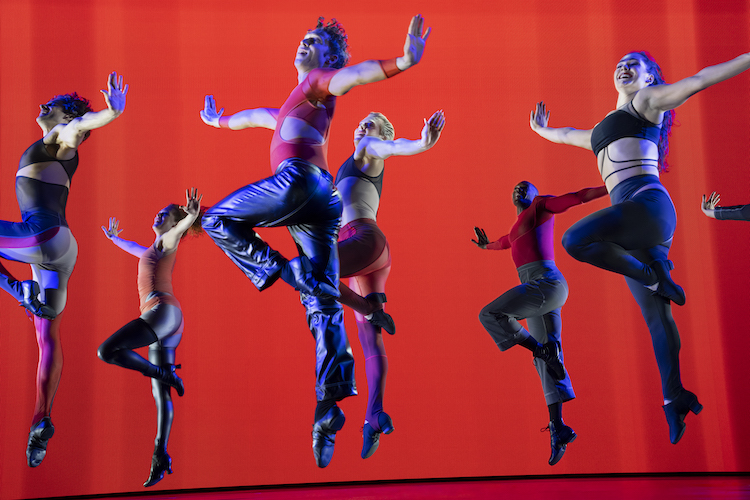 a chorus of dancers in a unison jump dressed in tight shiny clothing, everyone different, some with muliti colors, some with bare midriffs, all ecstatic, jumping against a red background