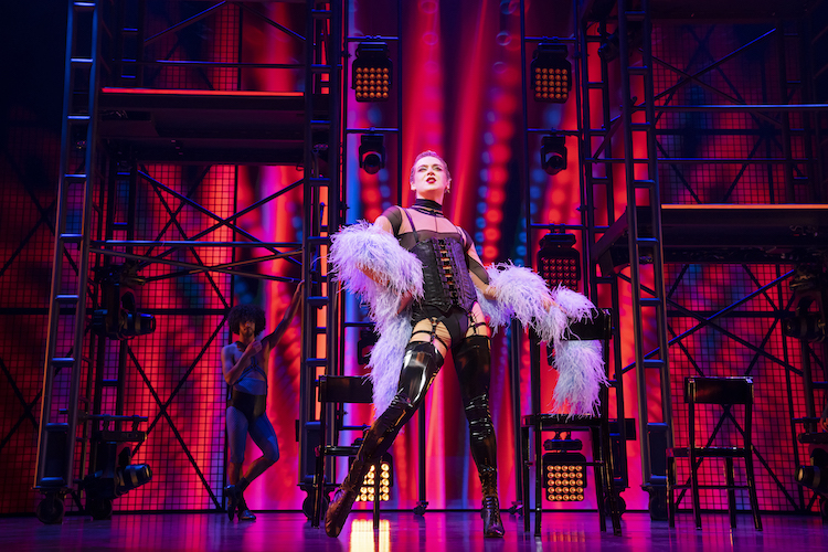 against scaffolding and a bright neon red backdrop a person with short slicked back hair a leather bustier with garters and they high black rubber heeled boots, stares with sass  and one hip cocked out at the audience. This person also rocks a wild feather boa that looks to be pastel purple.
