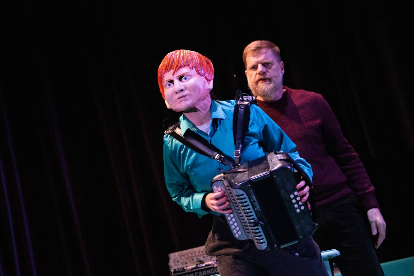 The masked dancer leans forward in front of the accordianist. His face full of concern.