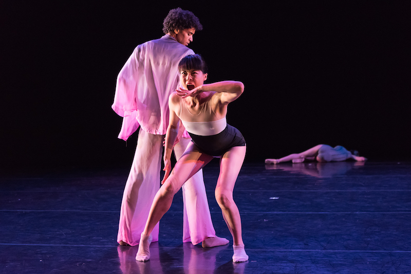 A women in a striped leotard bends at her waist and opens her mouth wide. She sticks her hand near her mouth as if she is going to bite it. A man watches her over his shoulder.