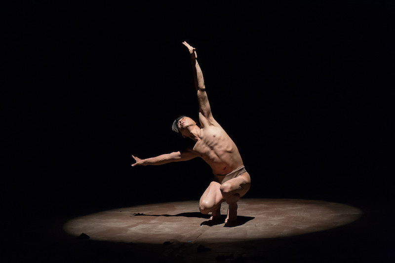 A man crouches down under a spotlight. His left arm extends to the ceiling