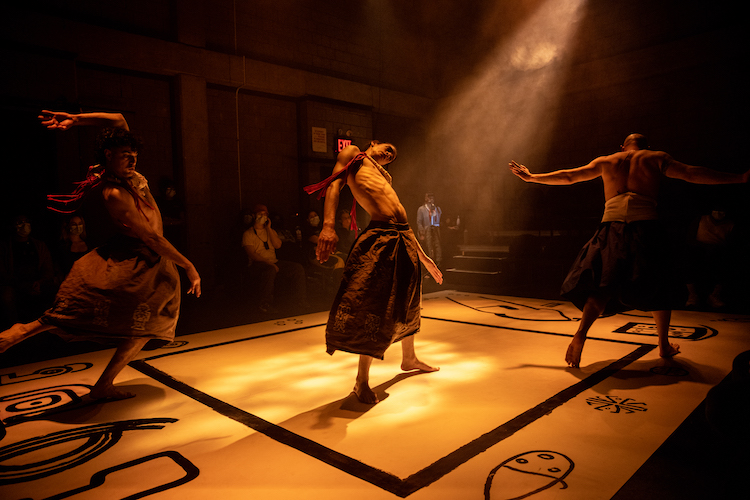 on a stage with a black square drawn in its center and surrounded by designs of petroglyphs , three men shirtless in skirts dance, each performing their own moves. We sense they are on the same journey but not moving in unison.