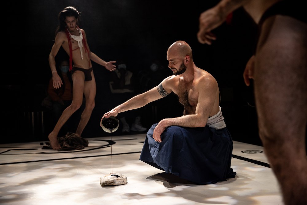 three male bodies in a ritual, in the center a shirtless bearded man in a blue skirt  pours a black liquid from a ceramic cup onto a stone. To his left and right are men wearing only dance belts and various colors of string about them. Each is immersed in their aspect of the ritual