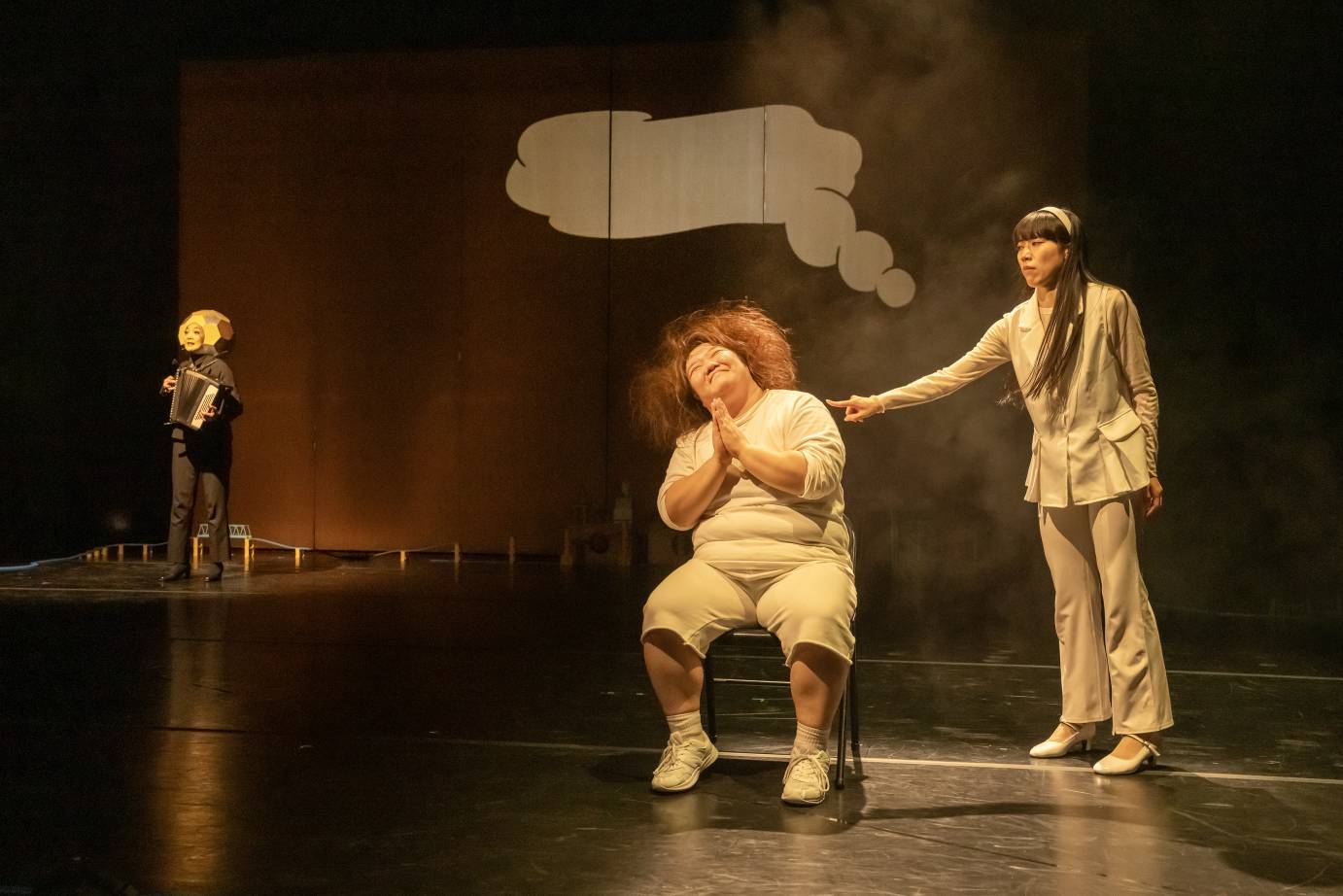 A beaming woman sits in a chair as a woman behind looks on pensively. The moon is standing far away to their right. A white 'speech bubble' is behind them blank.