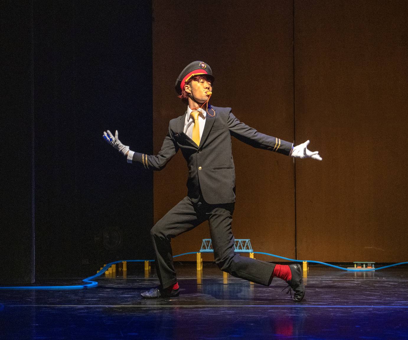A man wearing a gray suit, white glovers, red socks and conductor's cap blows a whistle as he towers over a miniature bridge.