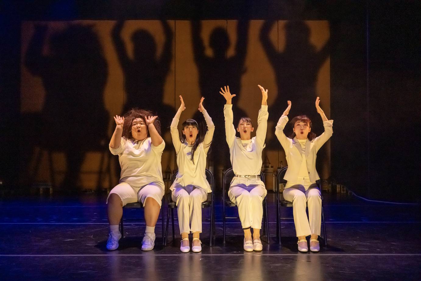 Four seated women, dressed in white with vertical arms above their heads, open their mouths as if reacting.