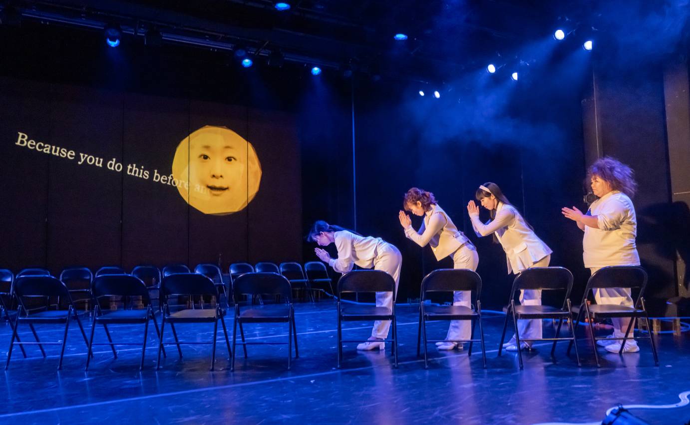 Four women in shite bow to a yellow moon face projected in front of them. Out of the moon's mouth come the written words, 
