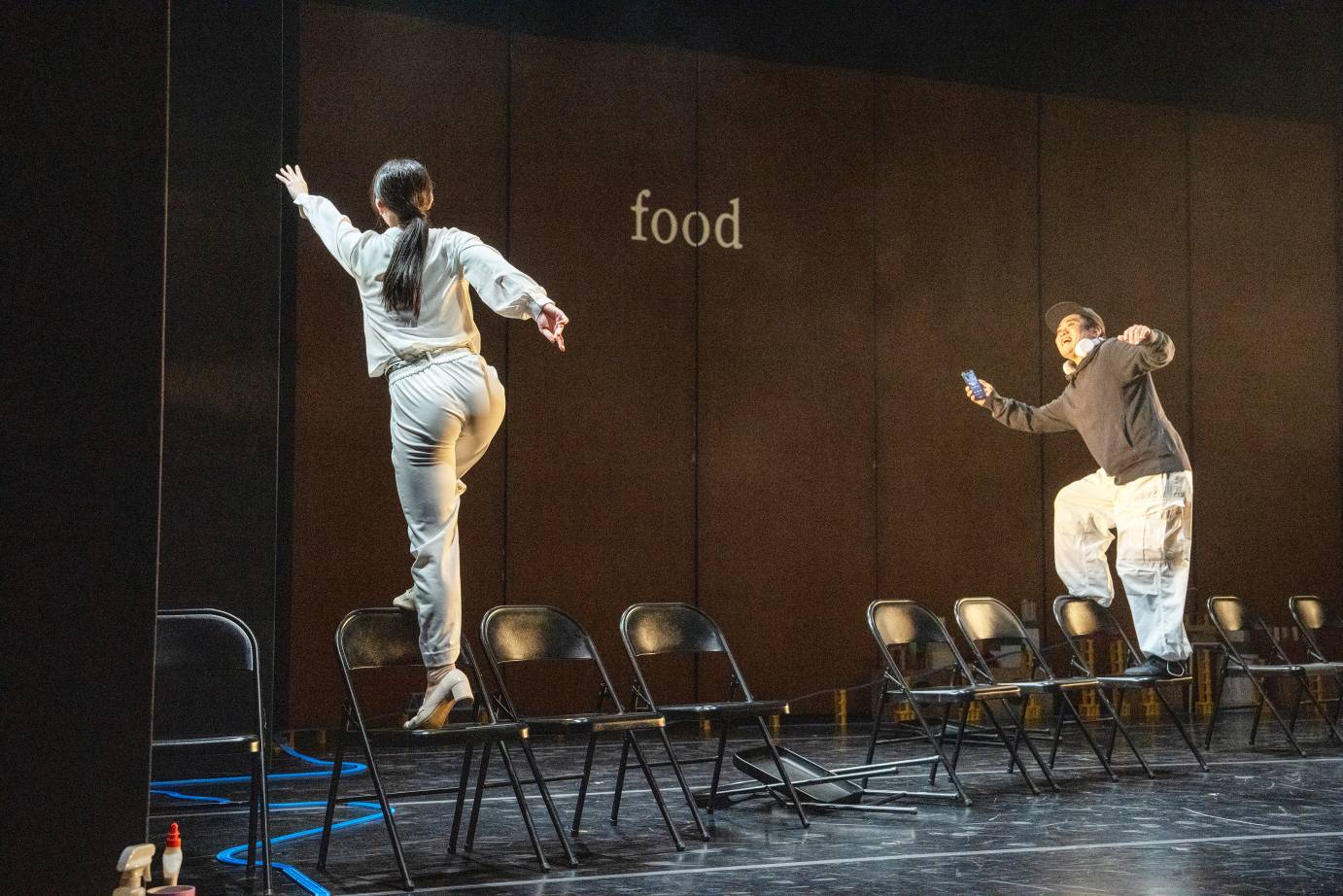 A woman in white stands on a black chair with her back to the audience. She reaches toward the upstage wall on which is projected the word, 
