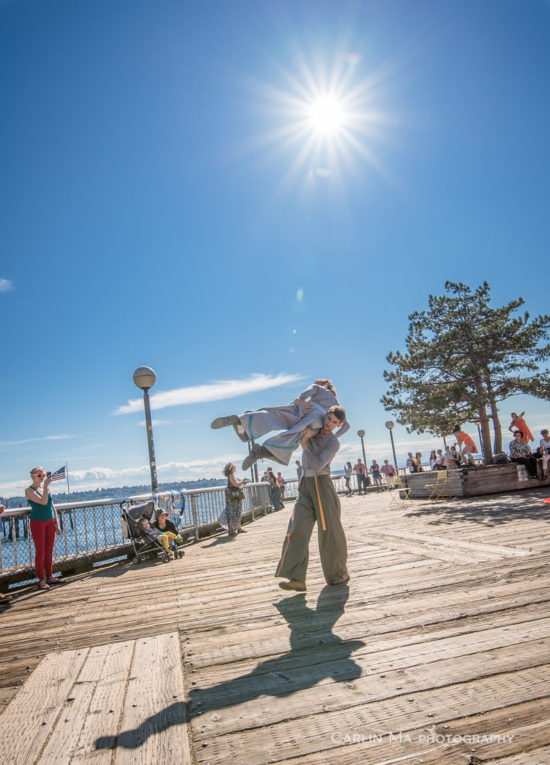 A man lifts a woman on a boardwalk under a beaming sun